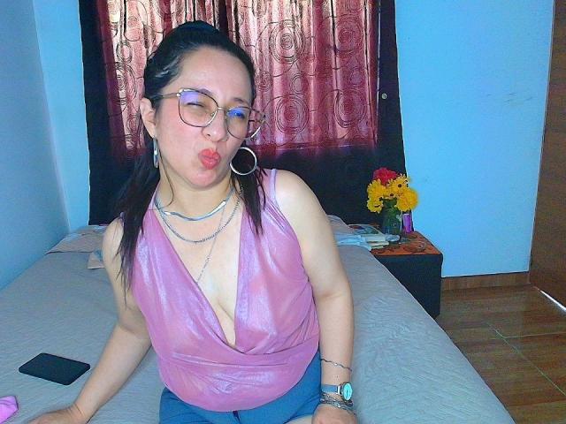VictoriaRousse with pouty lips, wearing a shiny pink sleeveless top, blue jeans, glasses, hoop earrings, and a watch. Background includes red curtains, flower vase, and bed. Bright lighting.