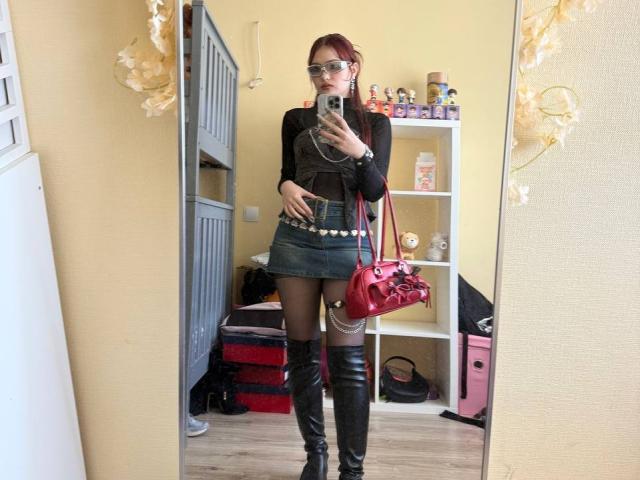 Bubochka posiert in einem Spiegel-Selfie und trägt ein schwarzes Oberteil, einen kurzen schwarzen Rock, einen mit Nieten besetzten Gürtel, schwarze hohe Stiefel und rote Handtasche. Sie schaut auf ihr Handy und strahlt eine selbstbewusste, sinnliche Ausstrahlung aus..