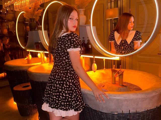 Bubochka in a black, short-sleeved, floral dress with white trim, standing in front of a glowing circular mirror and a lit bath tub, looking back over her shoulder. Warm, orange lighting.