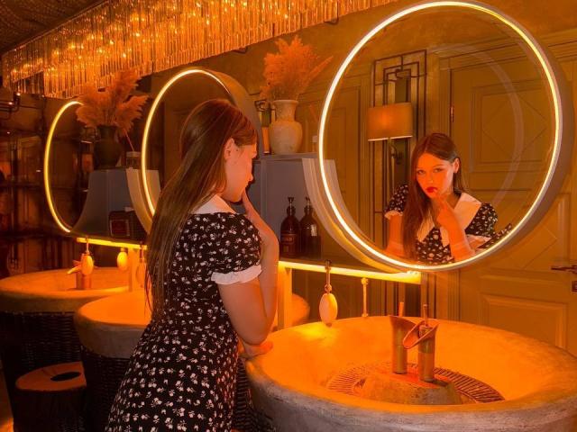 Bubochka, mit langem, schwarzem Haar und rotem Lippenstift, trägt ein schwarzes Kleid mit weißen Sternen und steht in einer schummrigen Bar. Sie betrachtete ihr Spiegelbild in einem runden Spiegel. Neonkreise leuchten hinter ihr.
.