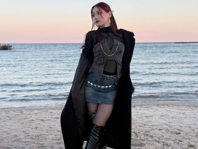 Bubochka steht an einem Strand, im Hintergrund das Meer, und trägt ein schwarzes Netzoberteil, einen Jeans-Miniröck, einen mit Nieten besetzten Gürtel und hohe Stiefel, die bis zu den Oberschenkeln reichen. Stiefel, schwarzer Mantel. Rote Haare, seitlicher Zopf, verführerischer Ausdruck.