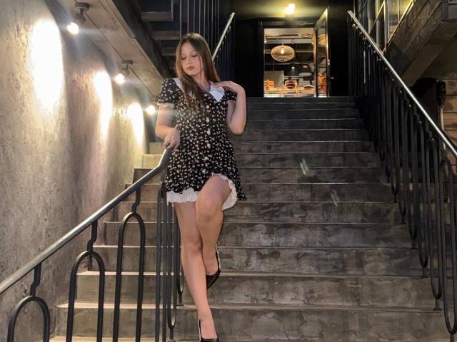 Bubochka, with long brown hair, stands on dimly lit concrete stairs, wearing a black polka-dot dress with white frills, one leg raised, revealing her thigh. Background lights create a sexy, provocative atmosphere.