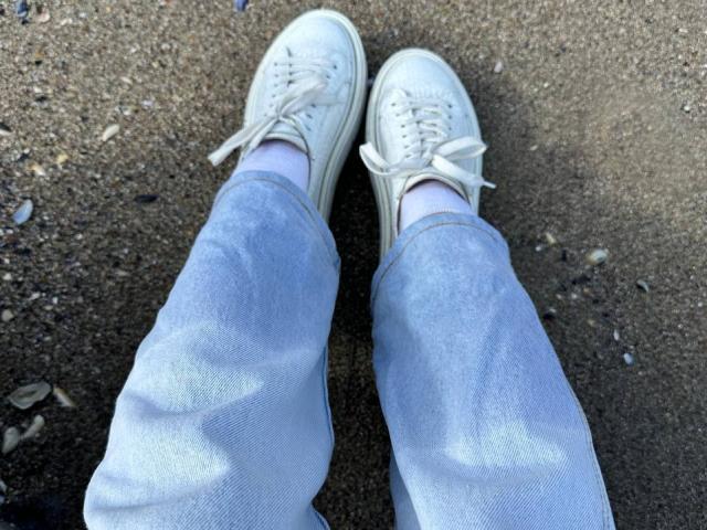 Close-up of Bubochka's feet, wearing white sneakers with white laces, and light blue, slightly wrinkled jeans, standing on rough, pebbly ground. Focused on the footwear and denim texture.