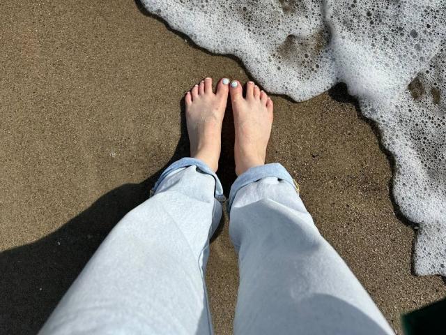 Nahaufnahme von Bubochkas nackten Füßen in weißen Hosen, die auf feuchtem Sand stehen, mit einer schaumigen Wasserlinie im Hintergrund. Die Füße stehen nah beieinander, die Zehen sind leicht gespreizt, und Sand klebt an den Füßen. Verdammt sexy, entspannte Strandatmosphäre..