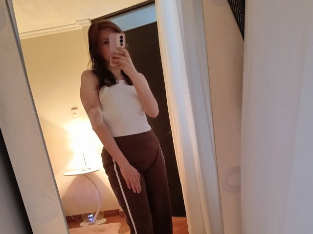 Selfie von KatyaRogersX in einem schlecht beleuchteten Raum, die ein weißes Top ohne Ärmel und schwarze Hosen trägt und ein Handy vor sich hält. ein Spiegel. Lampe und Bett sind im Hintergrund sichtbar. Sexuelle Spannung..