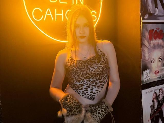 Bubochka steht in der Mitte, trägt ein Oberteil mit Leopardenmuster, ihre Hände sind in passenden Handschuhen, und im Hintergrund ist ein hellorangefarbenes Neon-Schild mit der Aufschrift "CACHES" zu sehen. Die rechte Seite zeigt Comic-Poster. Ein sanftes Licht betont ihre helle Haut..