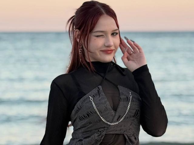 Fotografie von Bubochka mit dunkelrotem Haar, die ein schwarzes, durchsichtiges Oberteil und ein graues Kleid mit einer silbernen Kette trägt, stehend. vor einem unscharfen Hintergrund des Ozeans, lächelnd, mit einer Hand erhoben..