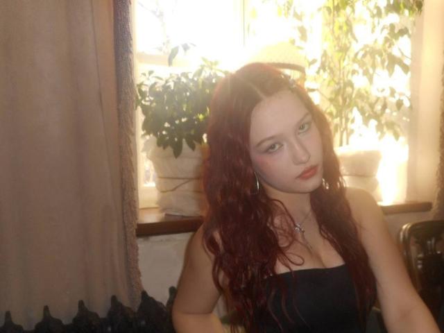 Photograph of a red-haired Bubochka with pale skin, wearing a black strapless top, sitting indoors by a bright window with sunlight filtering through leaves. She looks directly at the camera with a slightly bored expression.