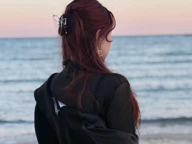 Bubochka mit langem, dunkelbraunem Haar im Pferdeschwanz, die ein schwarzes Oberteil trägt, steht mit dem Gesicht zum Meer gewandt. Sie ist von hinten beleuchtet, ihre Silhouette zeichnet sich deutlich vor den blauen Wellen und dem rosafarbenen Himmel ab. Verdammt heiß..