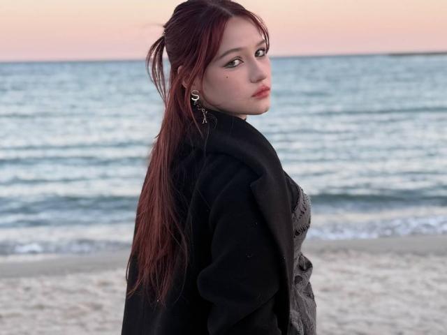 Photograph of a Bubochka with long red hair, wearing a black top, standing on a beach with ocean waves in the background. She looks back over her shoulder with a sultry expression.