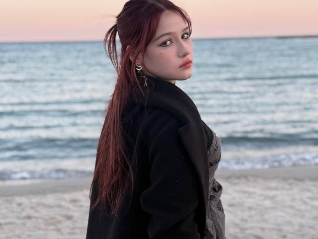 Bubochka with long red hair, pale skin, and black top, looking back over shoulder, ocean waves and sunset in background. Slight smirk, sexy vibe, casual beach setting.