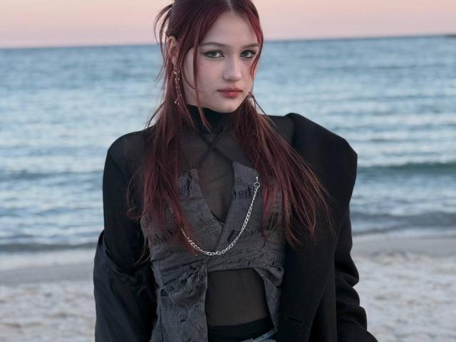 Bubochka mit langen, roten Haaren, heller Haut und einem schwarzen Kleid mit Schnürung steht am Strand. Sie trägt eine silberne Kette und blickt mit ernstem Gesichtsausdruck direkt in die Kamera. Meereswellen im Hintergrund.