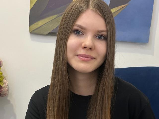 Photograph of a young woman, FireOfLovers, with long, straight brown hair, blue eyes, and pink lips, wearing a black shirt, against a white wall with abstract art and blue panel. Fucking gorgeous.