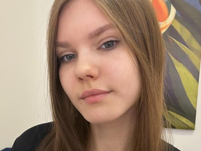 Portrait rapproché de FireOfLovers, avec une peau claire, des yeux bleus et des cheveux bruns lisses. Elle regarde directement l'appareil photo avec une légère moue, et elle porte un haut noir. Le fond comprend des formes abstraites vertes et oranges..