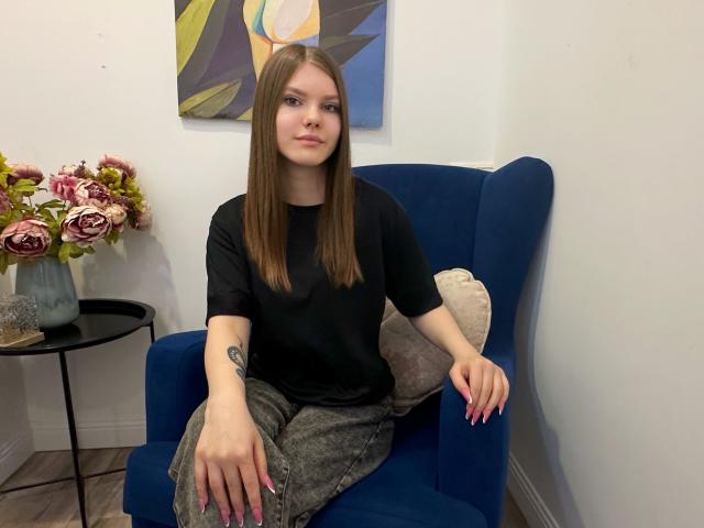 FireOfLovers, avec ses cheveux bruns lisses et sa peau pâle, est assise sur un fauteuil bleu, vêtue d'un haut noir et d'une jupe grise. Longues ongles, arrangement floral en arrière-plan, tension sexuelle..