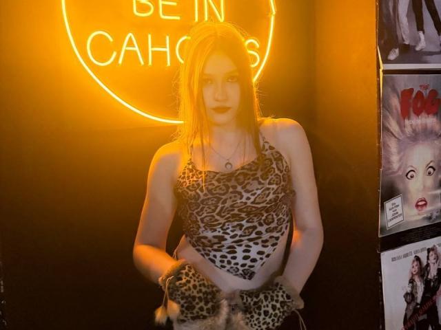Bubochka è in piedi di fronte a un cartello al neon con la scritta "BE IN CACHES", indossa una maglietta e dei pantaloni a stampa leopardo e tiene in mano tessuto con motivo leopardato. Poster con espressioni intense a destra. Incredibilmente sexy..