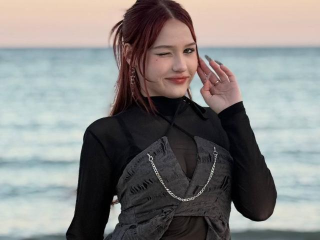 Fotografie von Bubochka, die am Meer steht und ein schwarzes, durchsichtiges, langärmliges Oberteil mit einem grauen, gemusterten detail im Bandstil und silberne Kette. Sie zwinkert, die Hand ist in der Nähe ihres Gesichts, sie sieht verführerisch aus..