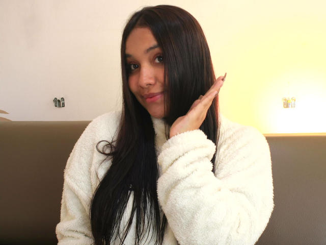 BelleLane, mit langem, schwarzem Haar, trägt einen weißen, flauschigen Pullover, sitzt auf einem braunen Sofa, ihre Hand berührt ihr Gesicht, und sie schaut... Direkt in die Kamera, leichtes Lächeln, sexuelle Spannung. Heller Hintergrund.