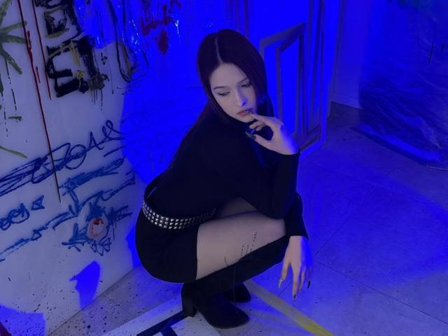 Bubochka, crouching in a dimly lit, graffiti-covered room, wears a black long-sleeve top and studded mini-skirt. Blue lighting casts a seductive glow, accentuating her sultry expression and pose.