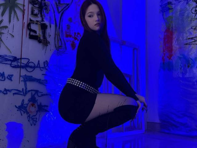 Bubochka, with dark hair and pale skin, crouches in a blue-lit room, wearing a black long-sleeve top, short black skirt with studded belt, and black thigh-high boots. Graffiti-covered wall behind.