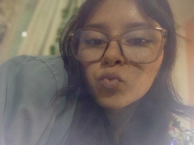 Close-up of EmiSchwarz with long black hair, wearing large brown glasses and a blue shirt. She's pouting with slightly pursed lips, looking directly at the camera. Sexual tension, cock hinted.