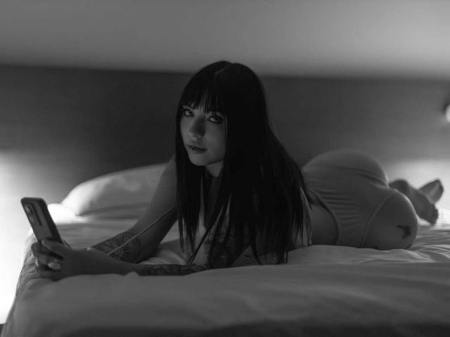 Black and white photo of KuroBabee, lying on a bed, face down, looking at a smartphone. She has long dark hair, tattooed arm, wearing tight shorts, legs raised, sexy vibe.