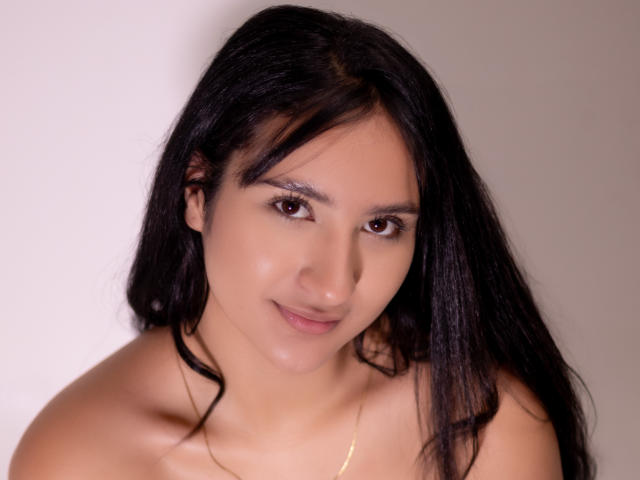 Photograph of ShinaJade with long black hair, fair skin, and brown eyes, smiling subtly. She's topless, showing soft shoulders, with a subtle sheen on her skin. Fucking attractive.