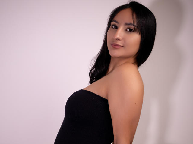 ShinaJade, com longos cabelos pretos, pele clara e olhos escuros, vestindo um vestido preto sem alças, vira-se ligeiramente para o lado. Olhando para a câmera com um sorriso discreto. Tensão sexual evidente..