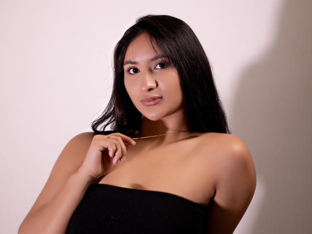 Photograph of ShinaJade, a sexy Asian woman with long black hair, wearing a strapless black top, looking directly at the camera with a slight smirk, hand near her chest. Soft lighting.