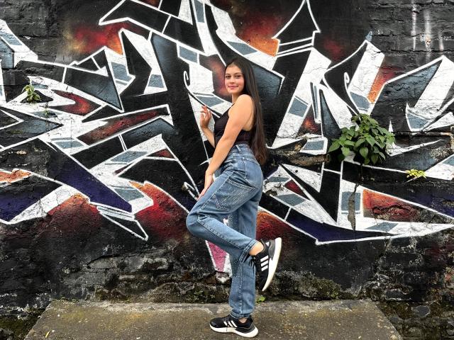 LizzaRose está de pé, com confiança, em frente a uma parede de grafite abstrata e áspera, com fragmentos coloridos e vibrantes. Ela está vestindo jeans azuis justos, uma camiseta branca e tênis pretos, e está sorrindo com um ar confiante..