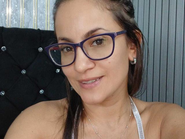 Primo piano di SolaraDubois, che indossa occhiali con montatura blu, un top a halter bianco e orecchini d'argento, con i capelli castani raccolti. Schiena scoperta, pelle chiara e un leggero sorriso, che trasmette un'aria sicura e sensuale..