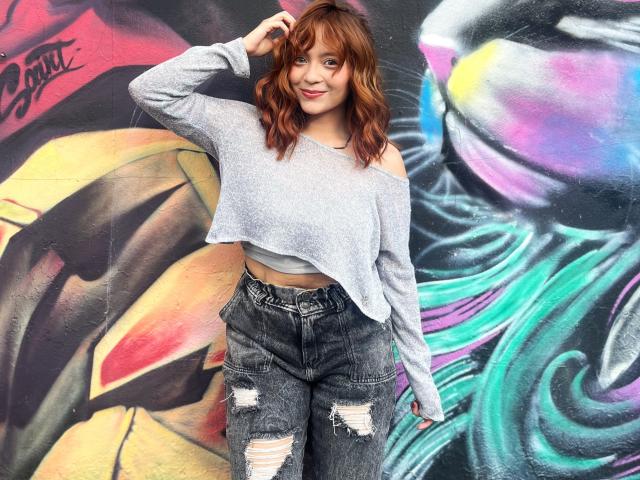 EmilyKlein, standing against a colorful graffiti wall, wears a white off-shoulder crop top and ripped black jeans. She has wavy brown hair, a relaxed pose, and a confident smile.