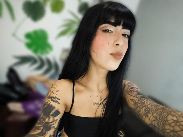 Fotografía de una joven, PeytonMorgan, con cabello largo y negro con flequillo, que lleva un top negro sin mangas, revelando una extensa tatuajes en sus brazos. Ella está mirando directamente a la cámara con una expresión seductora. Su piel es pálida y tiene pecas visibles. Sus tatuajes incluyen diseños florales y geométricos. Sus labios están ligeramente fruncidos. Las plantas de hojas verdes están borrosas en el fondo..