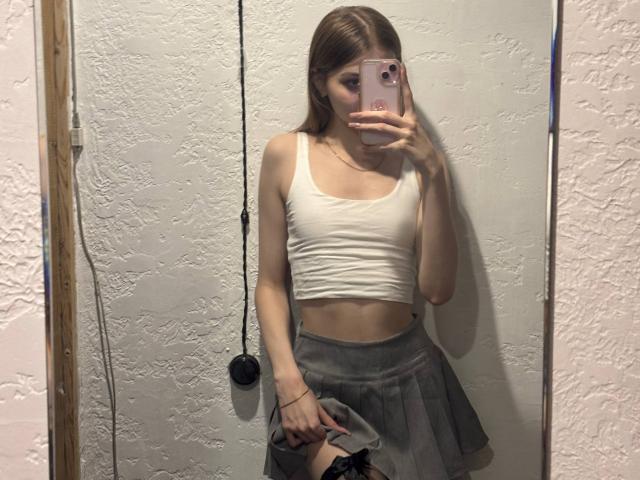 Photograph of CalistaSwiatek taking a mirror selfie, wearing a white crop top and gray pleated skirt, holding a phone with a pink case, slightly visible cock outline, casual pose.