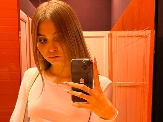 Fotografia di Calista Swiatek, in piedi in un bagno rosso ben illuminato, che si scatta un selfie allo specchio con un iPhone. Ha i capelli lunghi e castani, una camicia bianca e un'espressione intensa. La sua mano è visibile mentre tiene il telefono..