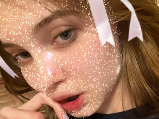 Close-up van het gezicht van Calista Swiatek, met glinsterende glitter die haar huid bedekt, bruine ogen die iets omhoog kijken, lippen gedeeld haar, witte linten in bruin haar, zacht licht, intiem, licht verleidelijke uitdrukking.