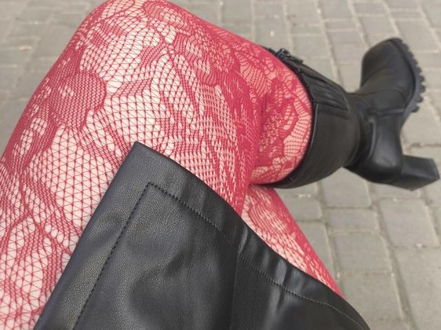 Close-up van Dreia's gekruiste benen, waarop rode netpanty's met een diamantpatroon en zwarte leren laarzen te zien zijn. De visnettextuur is duidelijk zichtbaar, met laarzen die lichtelijk versleten en gestructureerd zijn..