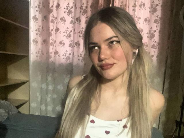 Photograph of RandaDaill, a young woman with long, straight blonde hair, wearing a white tank top with red heart patterns, looking slightly to the side with a subtle smile. Soft lighting.