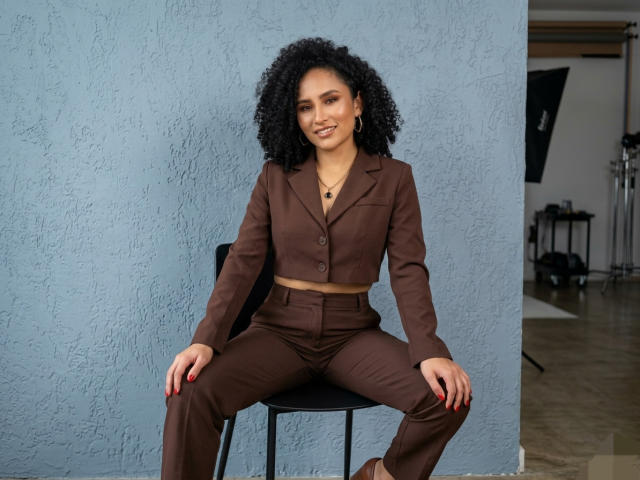 AbvrilDubois with curly black hair sits on a black stool against a textured blue wall, wearing a brown crop top and matching pants, legs spread, hands on thighs, smiling.
