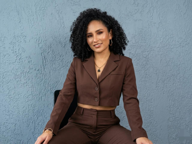 AbvrilDubois sits against a blue textured wall, wearing a brown crop top and high-waisted pants, smiling with curly black hair, hoop earrings, and a necklace. Fucking confident and sexy.