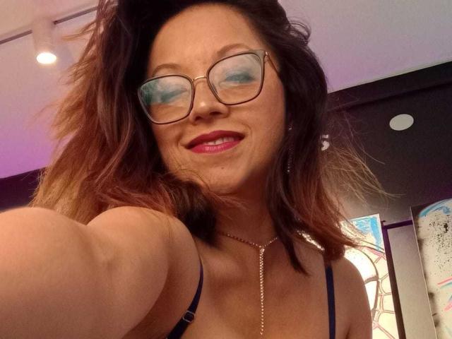 Foto ravvicinata di AngelinaBlanche con gli occhiali, rossetto rosso e capelli castani di lunghezza media, che allunga un braccio per selfie. Lei sta sorridendo, indossa una maglietta nera e una collana d'oro. C'è una tensione sessuale..