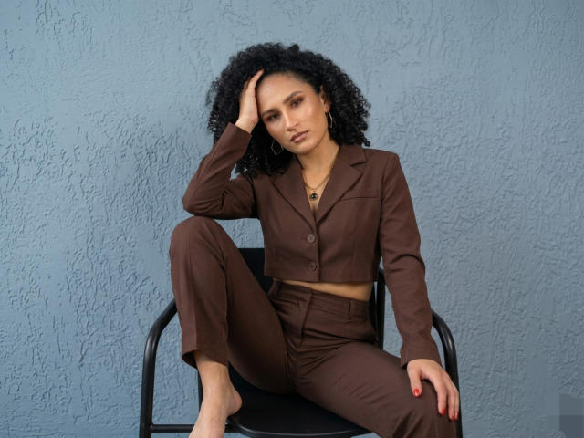 AbvrilDubois, com cabelos pretos encaracolados, está sentada em uma cadeira preta, vestindo uma blusa cropped e calças marrons, com a mão no cabelo e a perna levantada. Parede azul com textura, unhas vermelhas, expressão séria.