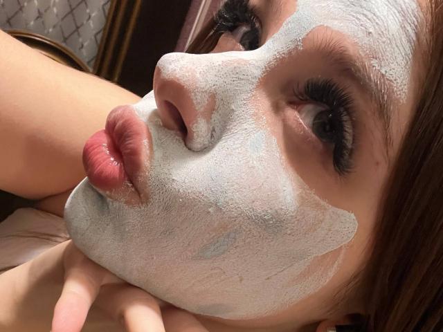 Close-up de NicolBonuz com uma máscara facial branca, lábios vermelhos curvados para baixo e olhos semicerrados. A luz do sol realça seus cílios escuros. Sua mão repousa em sua bochecha. Muito atraente..