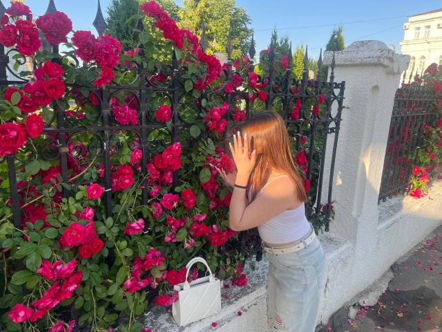 NicolBonuz, con el cabello castaño, mojado y de largo medio, se apoya contra una valla de hierro negro cubierta de vibrantes rosas rojas. Ella se cubre la cara con las manos, vistiendo un top blanco de tirantes y jeans de cintura alta. Bolso blanco en el suelo.