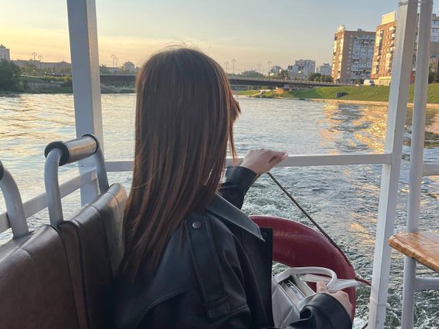 NicolBonuz è seduta su una piccola barca, con la schiena rivolta verso la telecamera, e osserva un tramonto sul fiume. Capelli lunghi, giacca nera, mano sul corrimano di una barca, edifici urbani in lontananza. Atmosfera al tramonto, semplicemente fantastica..