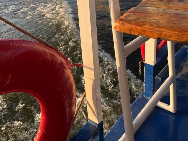 Close-up of a red life ring attached to a white and blue metal railing on a boat. Sunlight highlights the wood table on the right, with choppy water in the background.