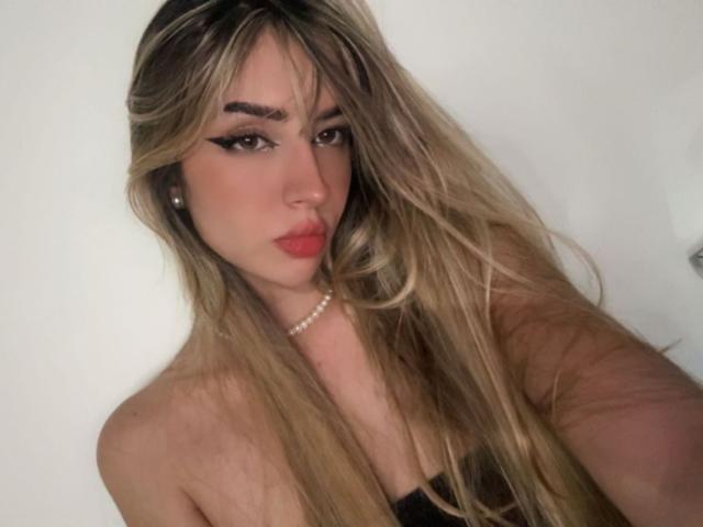Fotografie von Amelia Collins, mit langen, welligen blonden Haaren, starkem schwarzem Eyeliner, rotem Lippenstift, einer Perlenkette und Schwarz top ohne Träger. Sie hat einen verführerischen, intensiven Ausdruck und eine sexy Ausstrahlung..