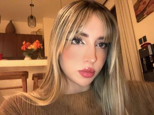 Close-up selfie of AmeliaCollins, with long blonde hair, heavy makeup, red lipstick, and large eyes. She wears a brown top, with a kitchen in the background, featuring red flowers. Fucking sexy.