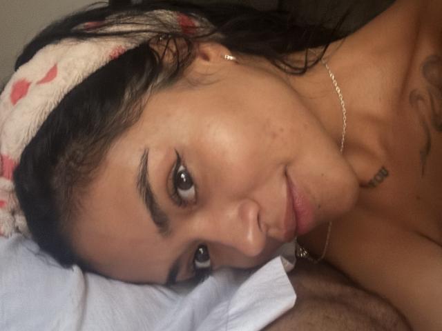 Gros plan de MajoJones, nue, allongée sur un oreiller blanc, la tête penchée, avec des cheveux foncés et un bandeau blanc et rouge. Petit tatouage sur le cou, regard séduisant, peau légèrement rosée..