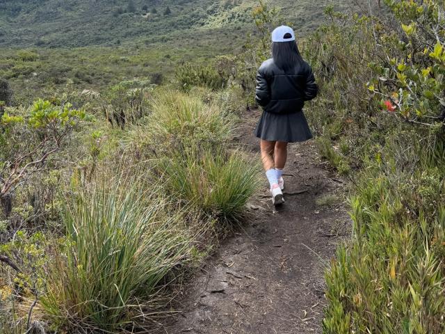 MajoJones, de espaldas, camina por un sendero de tierra a través de una vegetación densa. Llevando una chaqueta negra, una gorra blanca, una falda negra corta y calcetines blancos. La tensión sexual es evidente en su postura segura y sensual..